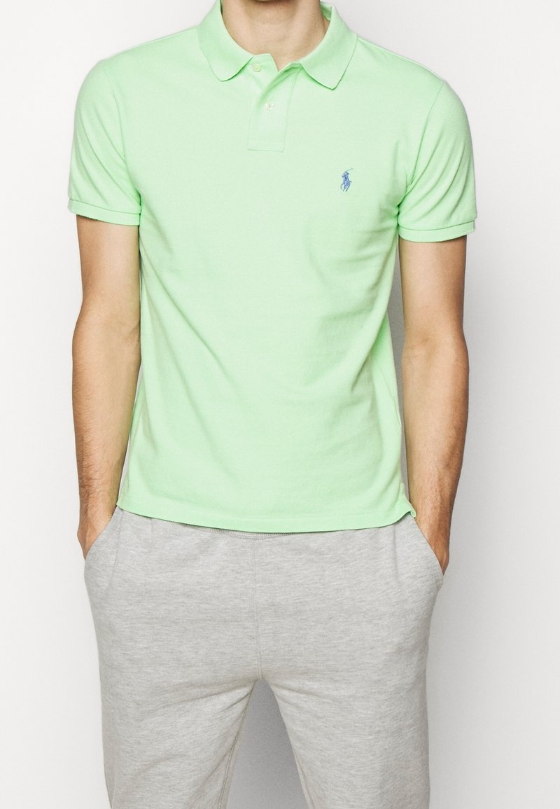 Muž v světle zeleném polo tričku s krátkým rukávem a malým vyšitým logem, světlé šedé tepláky, ruce v kapsách.