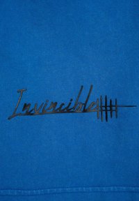 Niebieski bawełniany sweter z fakturowanym czarnym haftowanym logo z napisem "Invincible" i pięcioma pionowymi liniami poniżej.