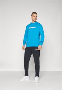 Niebieska bluza z kapturem z logo "HUMMEL", zestawiona z czarnymi spodniami dresowymi z pasującym brandingiem. Model nosi białe buty sportowe.