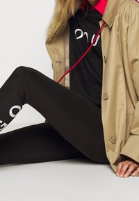 Trench beige sopra una camicia nera con dettagli rossi, abbinato a leggings neri con testo bianco stampato sulla parte inferiore della gamba.