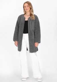 Cappotto grigio e peloso con collo a scialle, maniche lunghe e tasca sul petto con zip. Indossato sopra un top nero e abbinato a pantaloni bianchi.