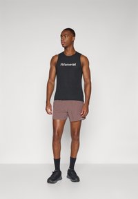 Sort ærmeløs tanktop med hvidt logo, brune shorts og sorte sneakers. Modellen står lige med hænderne ned langs siden foran en neutral baggrund.