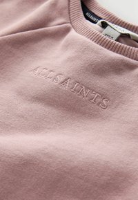 Tolmust roosa puuvillane särk ribakraega ja tõstetud tikitud "ALLSAINTS" logo rinnal, näidates toote silti krae sees.