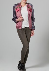 Chaqueta bomber de satén rosa con bordados florales, mangas grises, cuello de canalé y puños a rayas. Llevada sobre una camisa blanca con pantalones verde oliva.