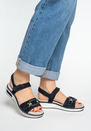 Feet wearing black open-toe sandals with gold buckle detail and red nail polish, paired with rolled-up blue jeans against a white background.