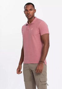 Rosa pikétröja med krage och tre knappar, med en broderad logotyp på vänster bröst, i kombination med ljusa kaki-byxor.