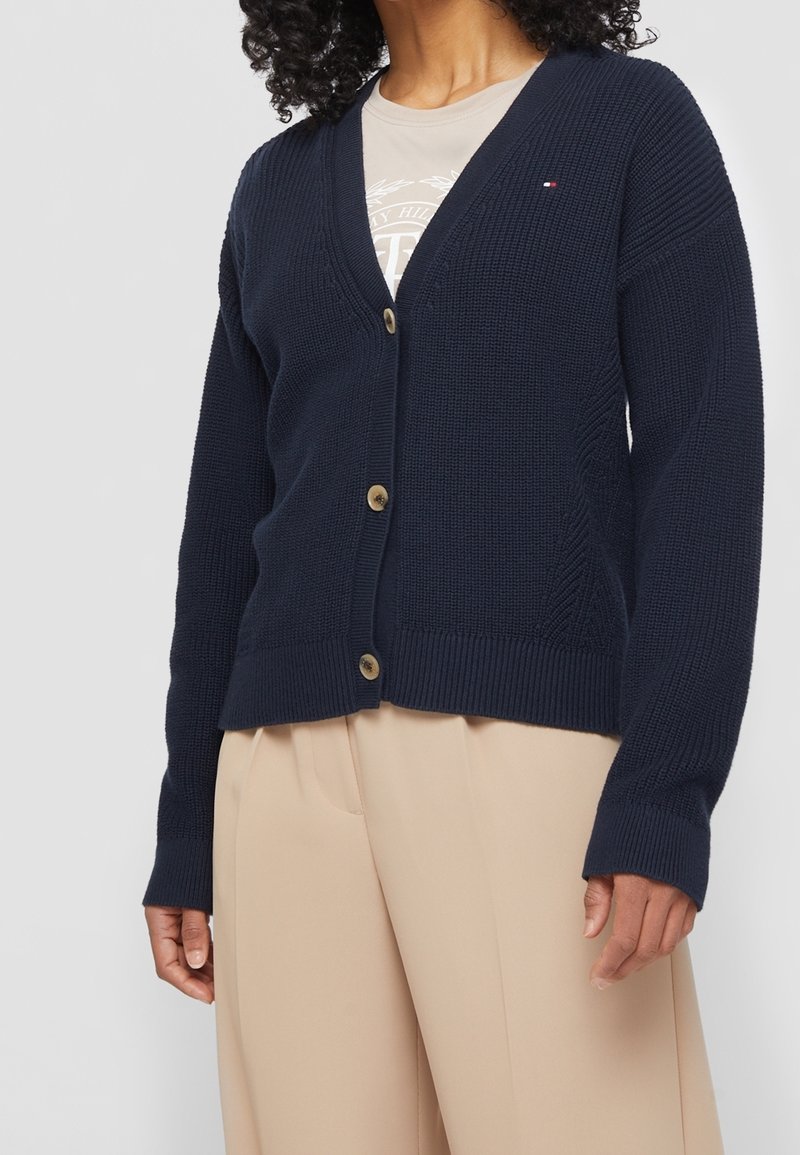 Marinblå stickad cardigan med v-ringning, prydd med gyllene knappar, ribbade ärmslut och ett texturerat mönster. Bärs över en ljusbeige skjorta.