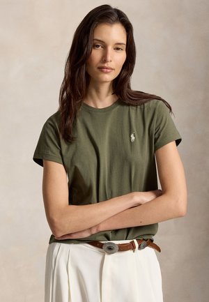 Femme aux longs cheveux bruns portant un t-shirt vert olive avec un petit logo et un pantalon blanc avec une ceinture marron, debout les bras croisés.
