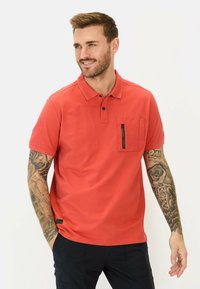Rode polo-shirt van gladde stof, met een kraag, korte mouwen en een ritszak op de borst. Aangevuld met een zwart etiket.