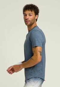 Camiseta azul de cuello redondo con una textura suave, que presenta mangas cortas y un dobladillo ligeramente curvado. El diseño incluye sutiles detalles de tatuaje en el brazo.