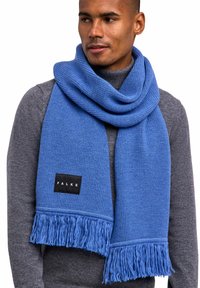 Écharpe tricotée bleue avec des franges aux extrémités et une étiquette noire avec "FALKE". Portée de manière décontractée sur un pull gris foncé.