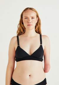 Γυναίκα με κόκκινα μαλλιά, φορώντας μαύρο σουτιέν και εσώρουχα, σε ποζάρει με φόντο απλής χρώματος, με μερικό αριστερό χέρι.