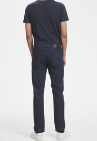 Pantalones de algodón mezclado en azul marino con corte recto, que cuentan con una etiqueta en la parte trasera de la cintura y dos bolsillos traseros, combinados con zapatillas blancas.