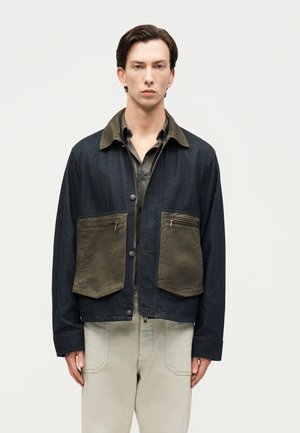 Άνδρας που φοράει σκούρο σακάκι με μεγάλες πράσινες τσέπες πάνω από ελιά πουκάμισο και ανοιχτόχρωμο παντελόνι, στέκεται μπροστά σε απλό λευκό φόντο.