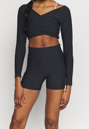 Vrouw met een zwart cropped top met lange mouwen over een roze sportbeha en zwarte, hoog geplaatste strakke korte broek, staand tegen een effen achtergrond.