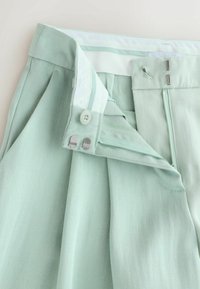 Light green trousers with an open zipper, button, and double hook-and-eye closure showing inner waistband details.