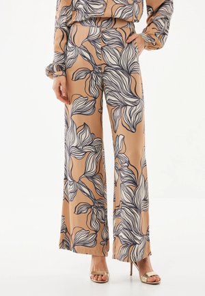 Beige broek met wijde pijpen en grote zwart-witte bloemenpatronen, gecombineerd met gouden open-toe hoge hakken en een bijpassende top.