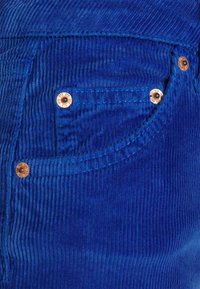 Tecido de corduroy azul com proeminentes costuras verticais, apresentando um bolso frontal acentuado por rebites de cobre e detalhes de costura contrastantes.