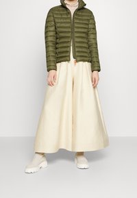 Olijfgroen pufferjack met horizontale stiksels, ritsluiting en hoge kraag. Gecombineerd met crèmekleurige broek met wijde pijpen en beige instappers.