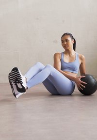 Világoskék sportmelltartó és hozzá illő leggings, fekete-fehér sportcipők, egy fekete medicinlabdát tart, sima padlón ül.