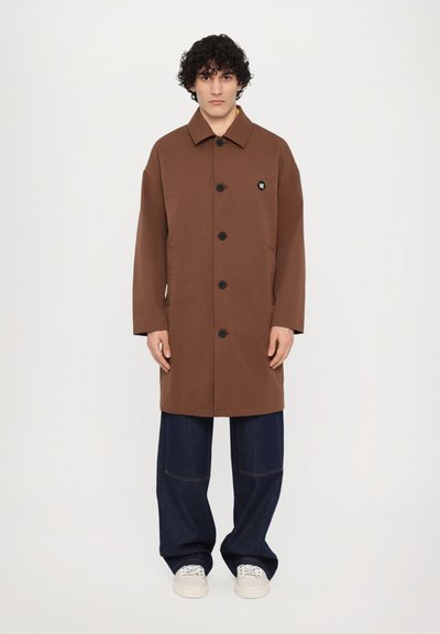 Abrigo marrón de doble botonadura con cuello, que presenta botones negros y un pequeño logo. Llevado con pantalones de mezclilla de pierna ancha y zapatos blancos.