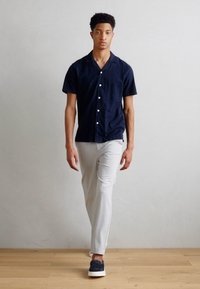 Camicetta navy a maniche corte con vestibilità rilassata, bottoni bianchi, pantaloni slim-fit grigio chiaro e scarpe scure su un pavimento di legno contro uno sfondo neutro.
