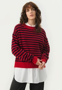 Pull rayé rouge et marine avec un col et des poignets en côte, porté par-dessus une chemise blanche à col, affichant une coupe décontractée et une texture douce.