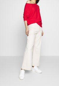 Maglione rosso a spalle scoperte, pantaloni a gamba larga color crema e sneakers bianche. Il tessuto sembra morbido e i pantaloni hanno un alto punto vita e tasche.