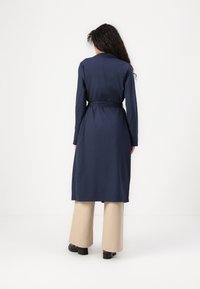 Navy blue dress with long sleeves, belt at the waist, and a flowing design, paired with beige wide-leg pants and black shoes.