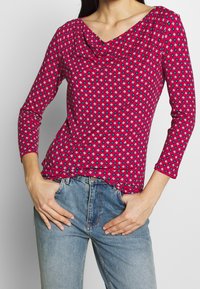 Figurpassad långärmad topp i livfull rosa med ett geometriskt mönster i blått och vitt. Har en muddkrage och en mjuk, elastisk textur.