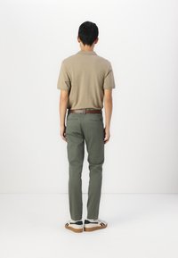 Polo shirt marron, pantalon vert olive, ceinture marron, baskets blanches et noires. Fond uni, vue de dos. Pas de motifs ou de caractéristiques visibles.