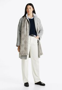 Gray wool coat with a high collar, navy sweater, white shirt, and white trousers. Scarf features a textured pattern and fringed edges. Black shoes.