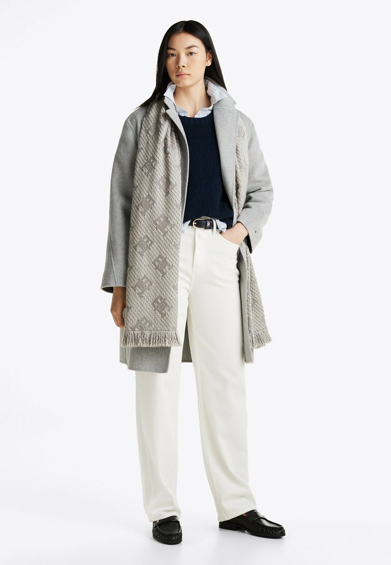 Gray wool coat with a high collar, navy sweater, white shirt, and white trousers. Scarf features a textured pattern and fringed edges. Black shoes.