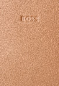 Brzoskwiniowa skóra o fakturowanym wykończeniu z tłoczonym logo "BOSS", podkreślającym jej gładką powierzchnię i subtelny połysk.