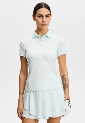 Kobieta o ciemnych włosach w jasnoniebieskim golfowym stroju, nosząca białą golfową rękawiczkę, stojąca o neutralnym wyrazie twarzy na jednolitym tle.