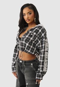 Przycięta czarno-biała bluza z kapturem w kratę, związywana sznurkiem, z zamkiem z przodu i logo na rękawie; zestawiona z ciemnymi dżinsowymi spodniami cargo z kieszeniami.