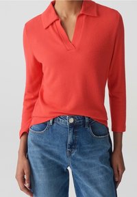 Haut corail à manches longues avec encolure en V et col. Porté avec un jean en denim bleu. Le tissu semble doux avec une finition texturée.