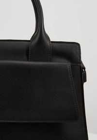Sac à main en cuir noir avec une poignée supérieure et une poche avant à rabat, présenté sur un fond clair uni.
