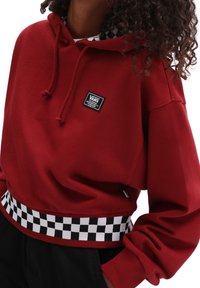 Persona que lleva una sudadera roja de Vans con un patrón de tablero de ajedrez en blanco y negro en la cinturilla y un parche con el logo de Vans en el pecho.