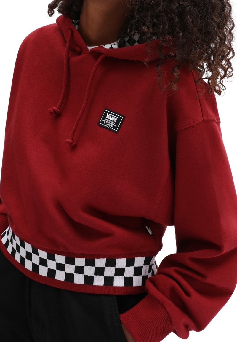 Persona que lleva una sudadera roja de Vans con un patrón de tablero de ajedrez en blanco y negro en la cinturilla y un parche con el logo de Vans en el pecho.