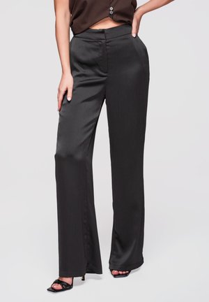 Kobieta nosząca czarne satynowe spodnie z szerokimi nogawkami i wysokim stanem, zestawione z czarnymi sandałami na obcasie, pozująca z jedną ręką na biodrze, a drugą w spoczynku.