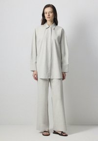 Chemise boutonnée oversize gris clair avec col et manches longues, associée à un pantalon large en tissu assorti et des sandales noires.