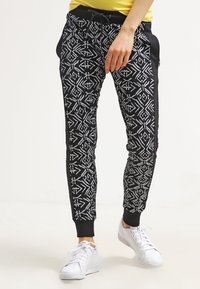 Zwarte joggers met een wit geometrisch patroon, elastische tailleband en zijzakken. Gecombineerd met witte sneakers en een geel topje.