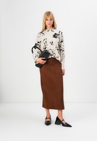 Donna che indossa una blusa lunga a maniche lunghe floreale beige, una gonna midi marrone, scarpe Mary Jane nere con il tacco e tiene in mano una borsa nera.