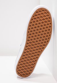 Semelle d'une basket blanche avec une semelle extérieure en caoutchouc marron à motif gaufré, inclinée contre un coin de mur blanc.