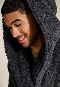 Donkergrijze, pluche badjas met kap, gemaakt van een getextureerde, pluizige stof, voorzien van een brede kraag en diepe kap die om de schouders van het model valt.
