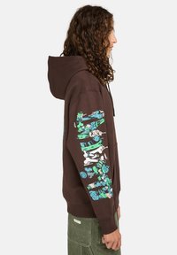 Bruine hoodie met een kangoeroezak en een trekkoordkap. Grafisch ontwerp in groen en blauw op de linkermouw. Comfortabele pasvorm.
