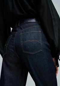 Persona che indossa jeans Salsa di un blu scuro con cuciture visibili sulle tasche posteriori e una maglietta nera a maniche lunghe su uno sfondo semplice.