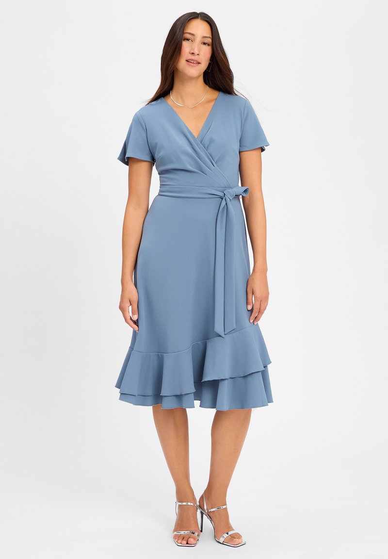 Hellblaues Wickelkleid mit kurzen Ärmeln, Taillengürtel und geschichtetem Rüschensaum. Glattes Material mit weicher Textur. Silberschimmernde Riemchensandalen.