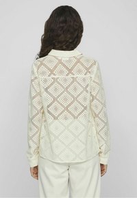 Blouse en dentelle crème clair avec motifs géométriques en forme de losange. Manches longues, poignets boutonnés et col classique. Détail transparent dans le dos.